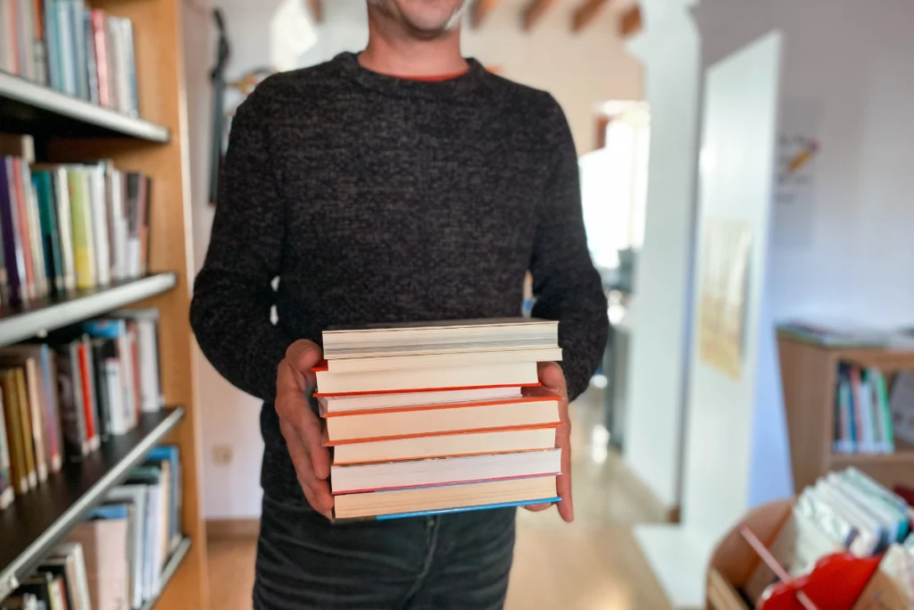 A photo of a many who is standing in a library aisle while holding a stack of books.
